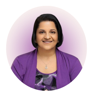 A woman with straight dark hair smiles, wearing a purple patterned top and matching purple cardigan, with a silver heart-shaped necklace, in front of a soft purple gradient background.